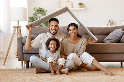 Shot of parents with child smiling and keeping roof mockup over heads while sitting on floor
